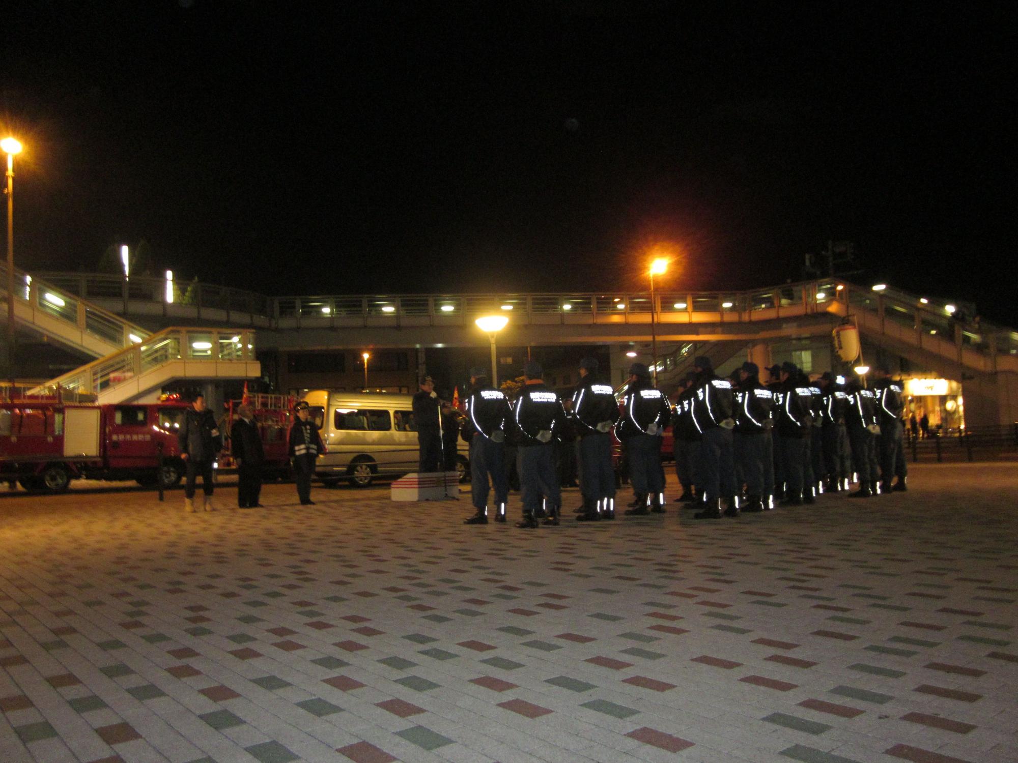 歩道橋がある夜の広場に、反射材のついた制服を着た消防団員が整列しており、高橋市長が団員に向けて演説している写真