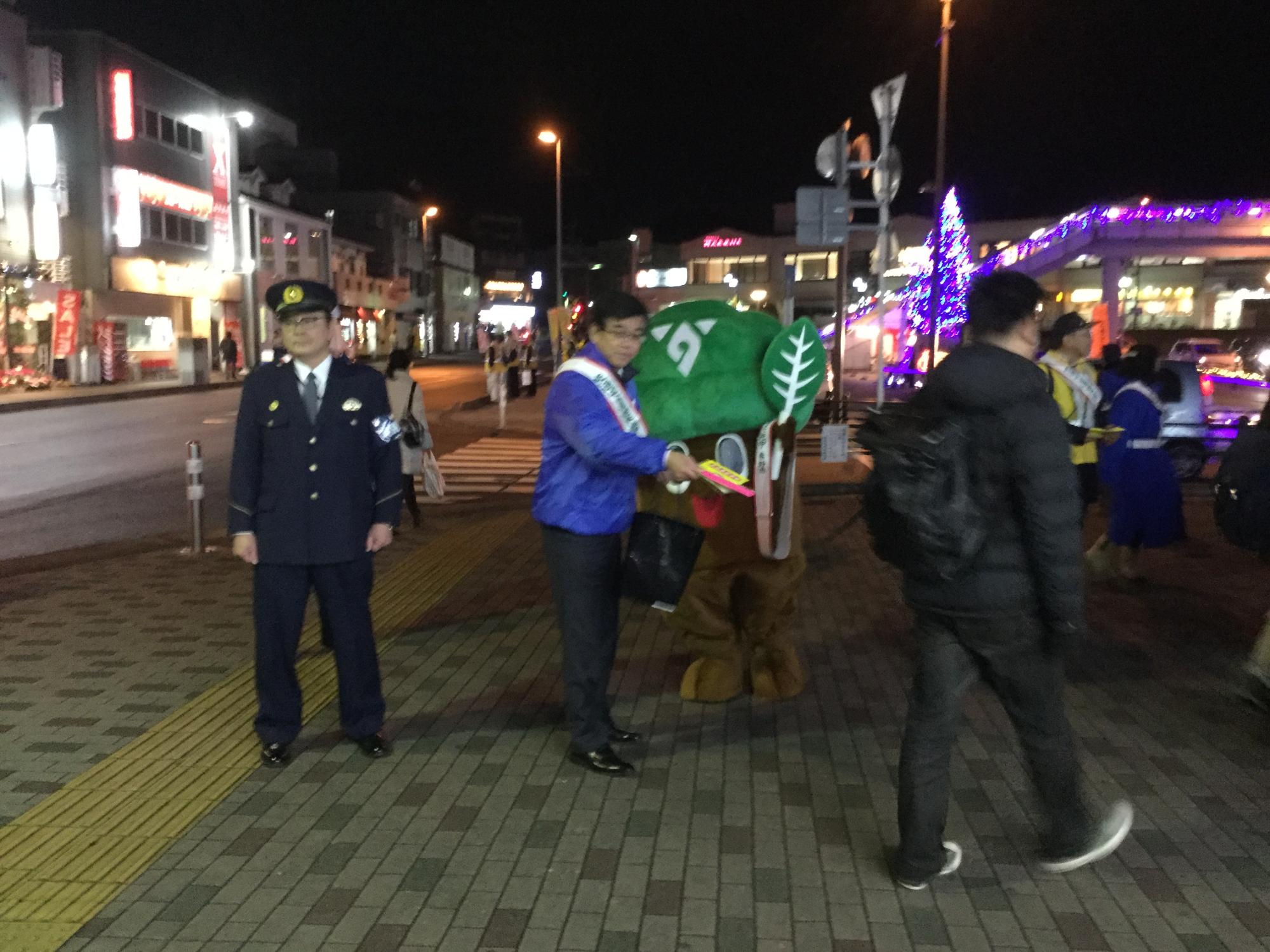 イルミネーションが光る夜の駅前広場で、襷を掛けた高橋市長ともりりんが、交通安全の呼びかけを行っている写真