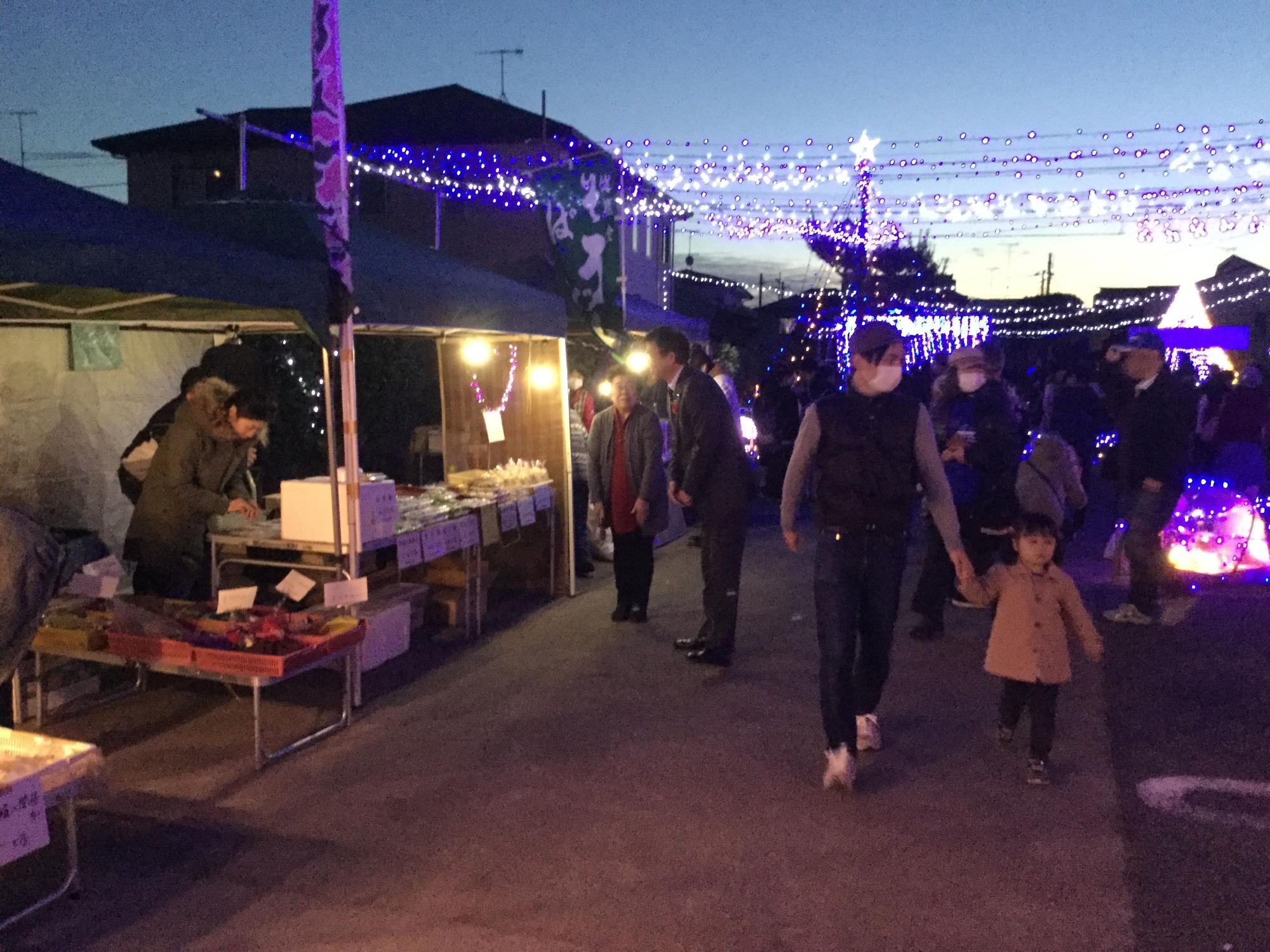 イルミネーションが輝く公園に屋台が並び、多くの人が行き交う中、手をつないだ親子が歩いている写真