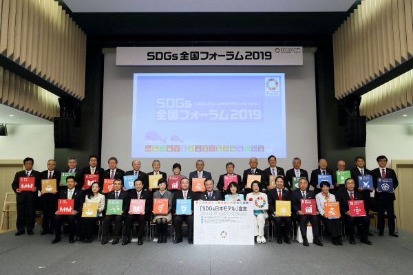 大きなスクリーンの前で、SDGsのロゴが入ったパネルやポスターを手に持ち記念撮影をしている集合写真