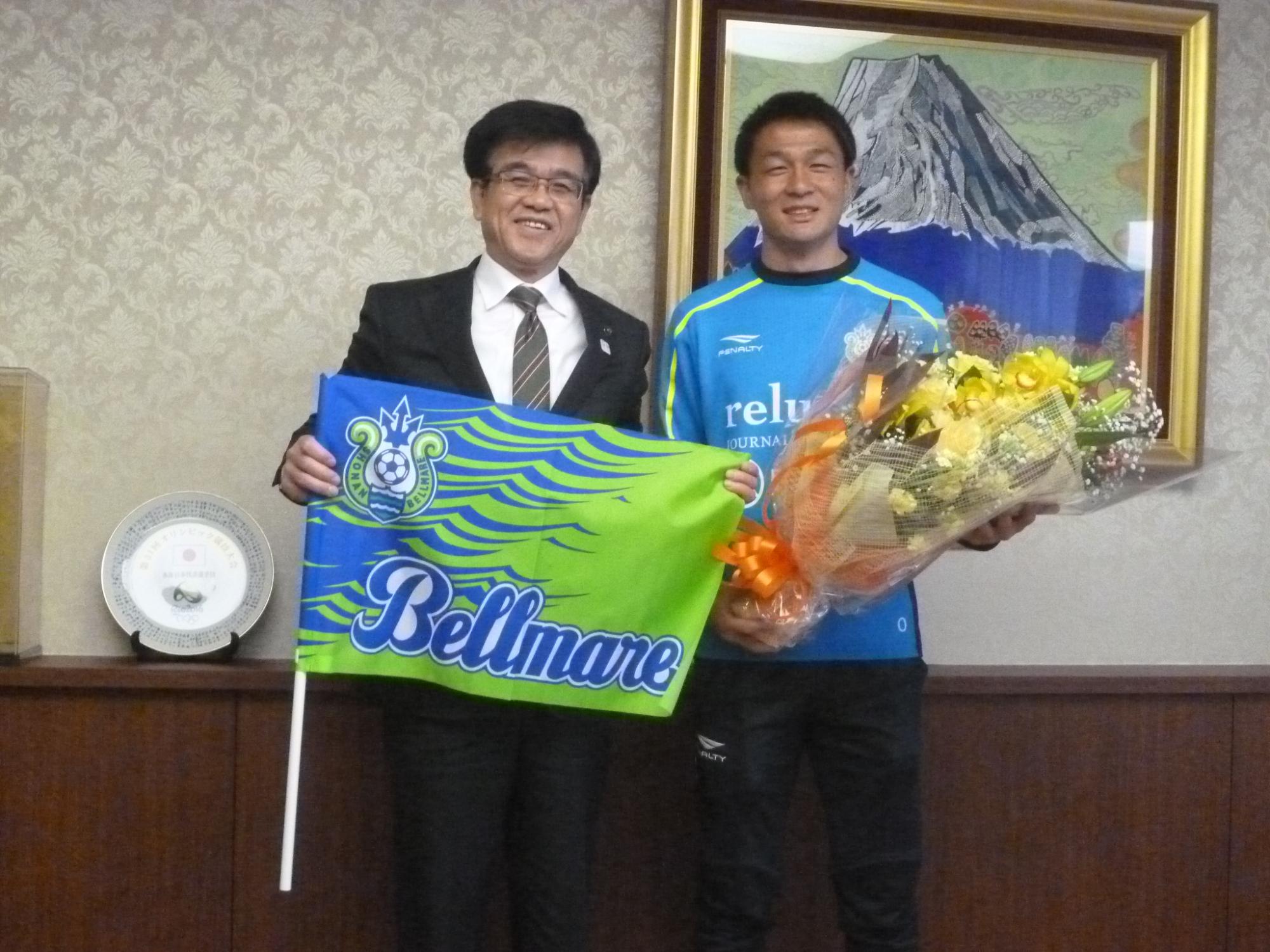 「Bellmare」と書かれた緑と青の旗を持った高橋市長と、花束を抱えた水色のユニフォーム姿の岡大暉選手が、並んで記念撮影をしている写真