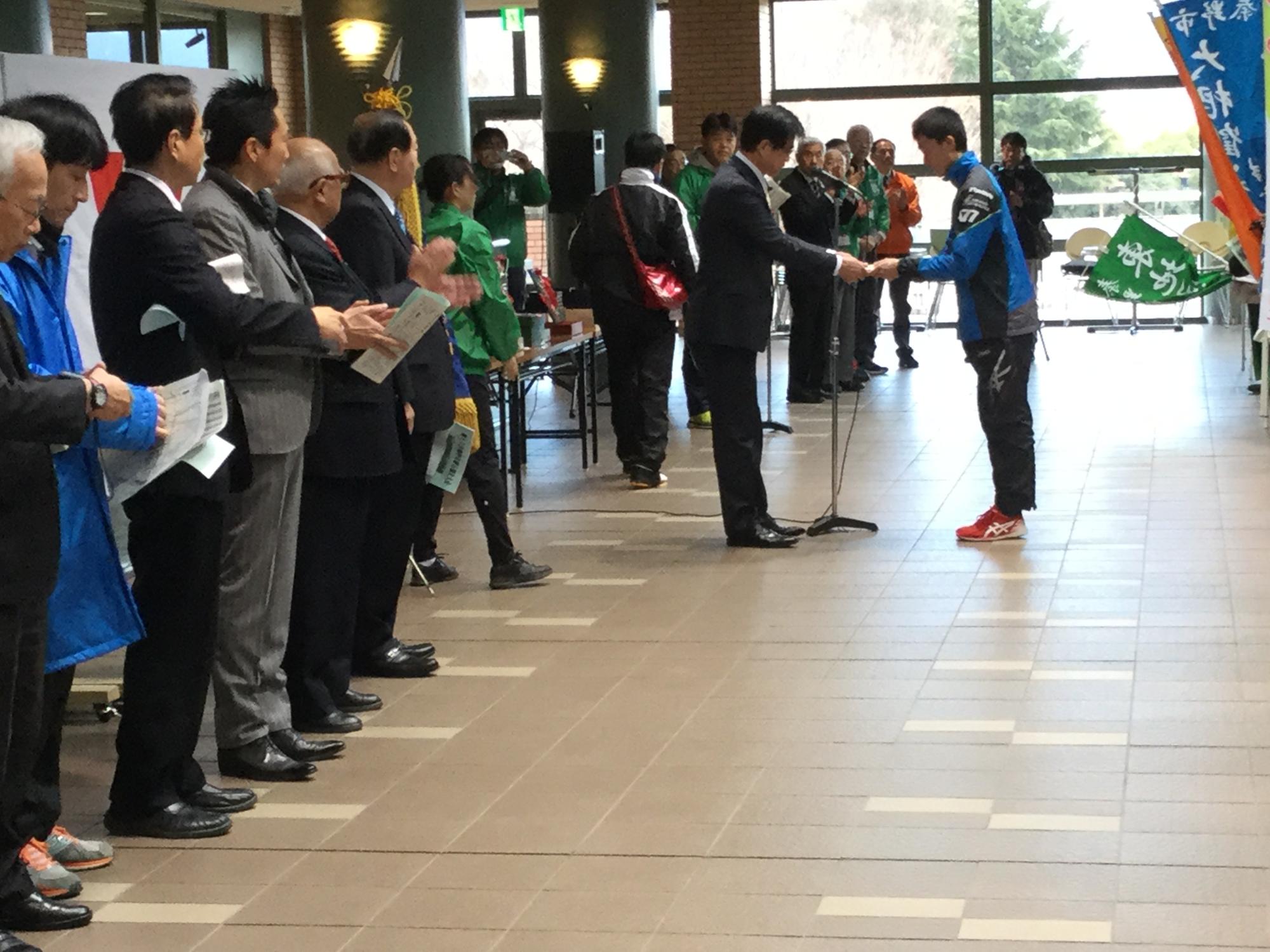 広いロビーのような屋内で、スポーツウエアを着た男性と高橋市長が向かい合っており、関係者が拍手を送っている写真