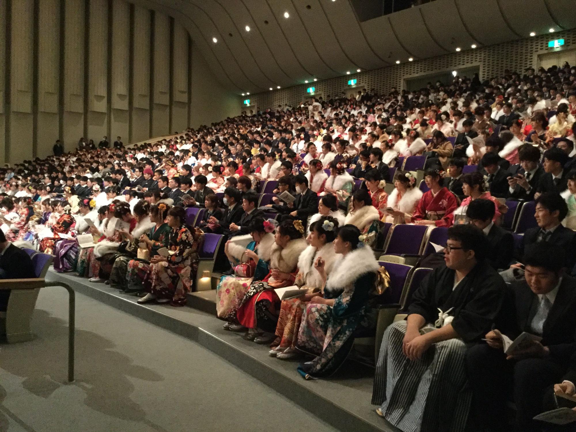 華やかな振袖や袴、スーツを着用した新成人が、ホール内の客席に座っている写真