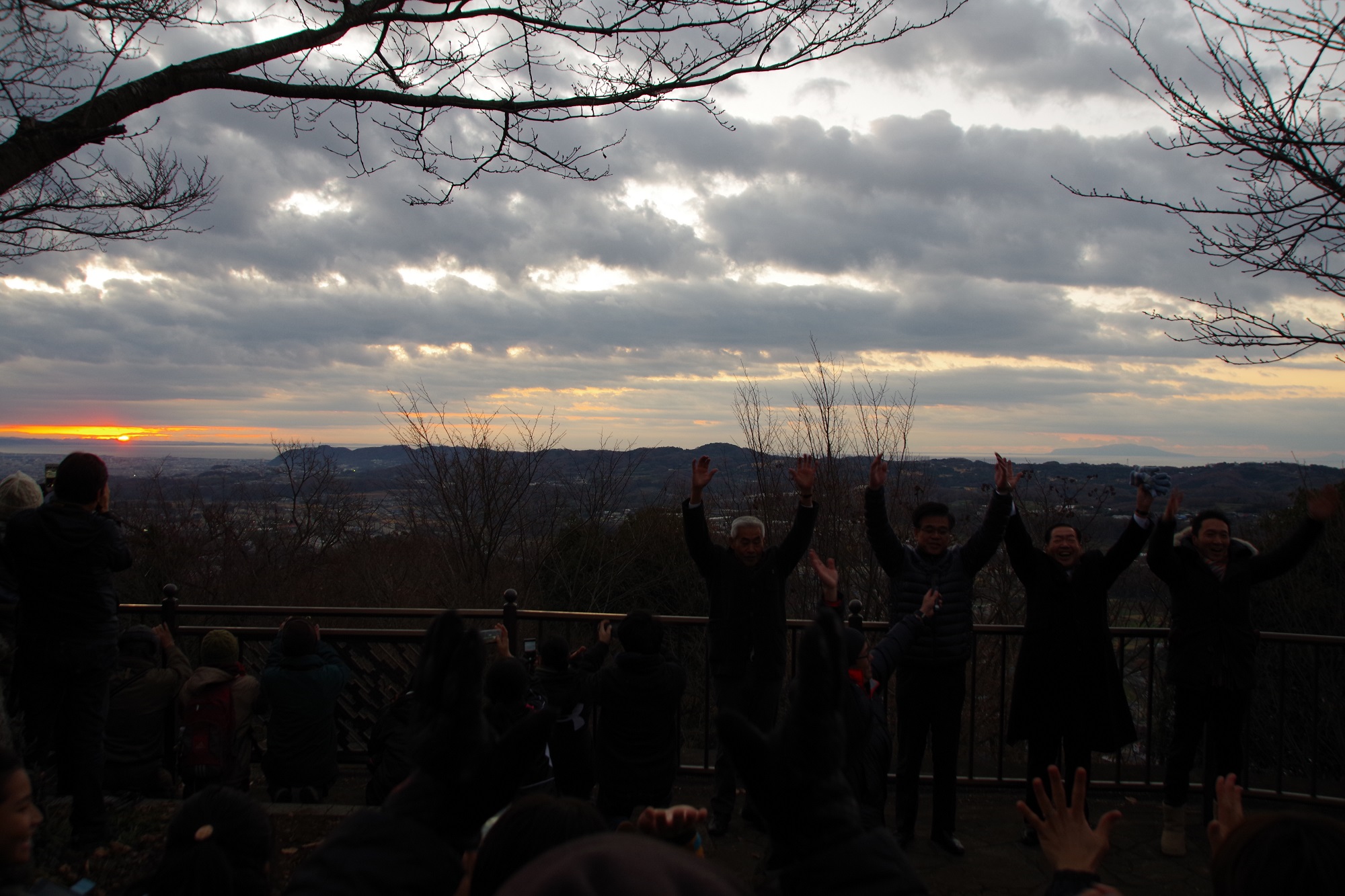 薄暗い元旦の早朝に、オレンジ色の日の出が現れ、高橋市長と参加者が両手をあげている様子の写真