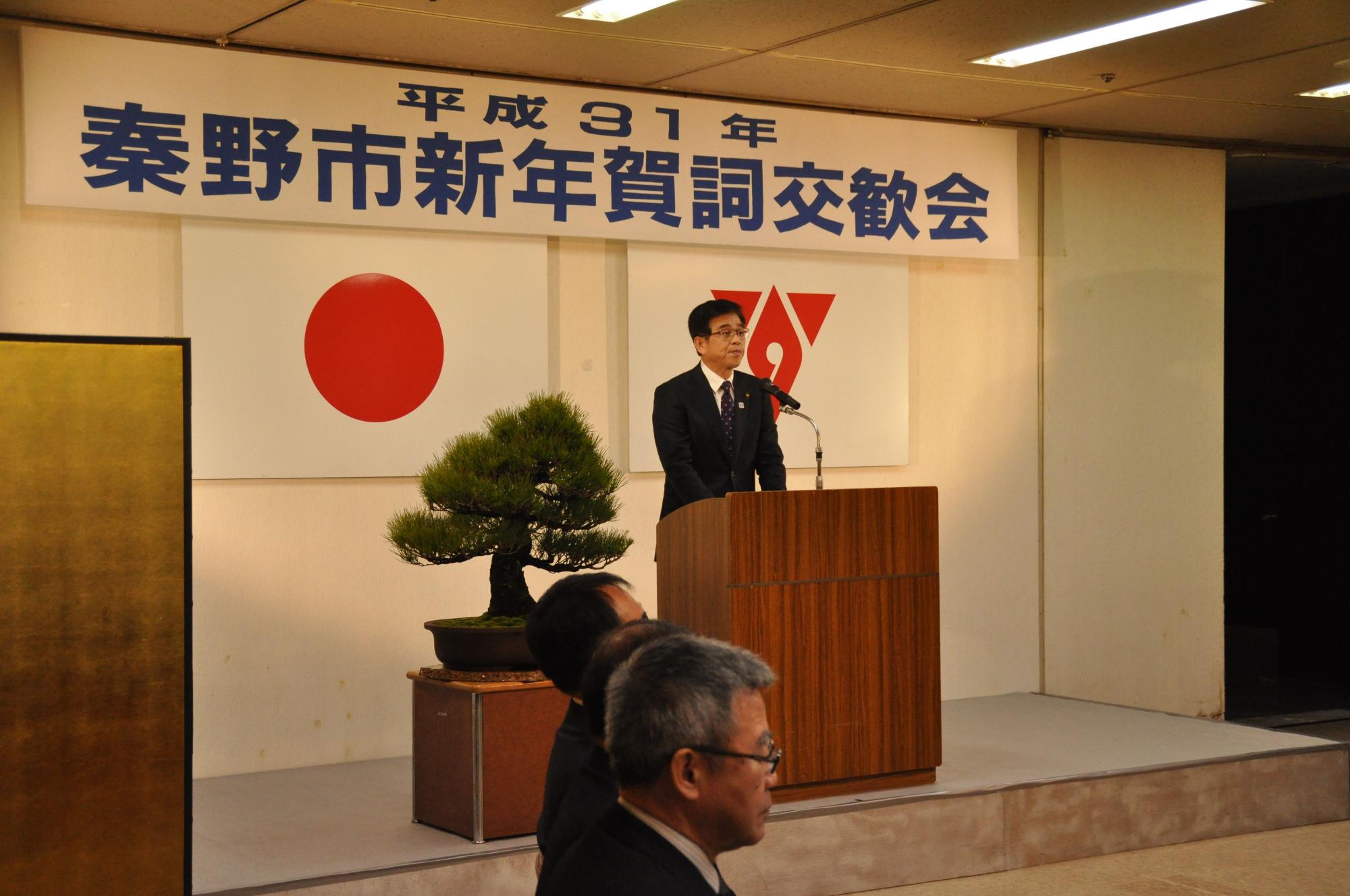 「秦野市新年賀詞交歓会」と書かれた横断幕がある会場で、檀上に立ち演説する高橋市長の写真
