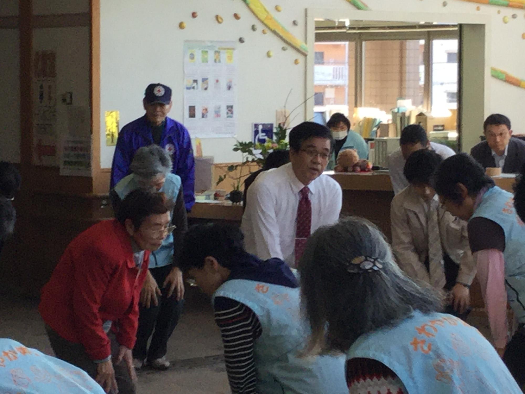 保健福祉センターの室内で、高橋市長と参加者達が膝に手を置き、前かがみになって体操をしている様子の写真