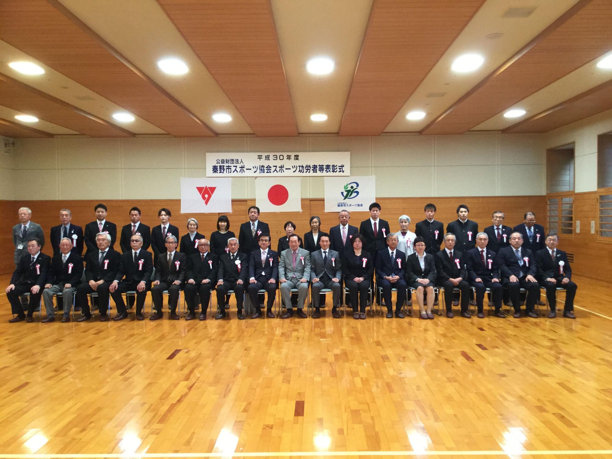 広い体育館に「秦野市スポーツ功労者等表彰式」の横断幕が掲げられており、正装した男女の功労者が整列した記念写真