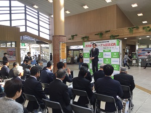 光が差し込む駅構内のような広い場所で、黒いスーツ姿の市長が「リニューアル記念式典」と書かれた緑色の看板の前で聴衆に向かって挨拶をしている様子の写真