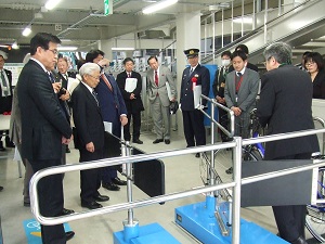 明るい屋内施設で、スーツ姿の市長と複数の男性や制服姿の警察官などが集まり、青い自転車ラックのような設備を囲んで説明を聞いている様子の写真
