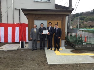 木目調の外壁を持つ公衆トイレの前に、スーツ姿の市長と関係者が並んだ除幕式の記念写真