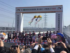 道路開通を祝う式典で、白い看板の下に並んだスーツ姿の関係者が、金色の玉と紅白や緑色のテープのついた紐に手を伸ばしている様子を、大勢の見物人がスマートフォンなどを構えている前景とともに捉えた写真