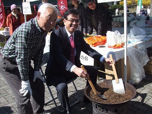 笑顔の市長と男性が屋外のイベントで、七輪に乗せられた大きな鍋の中の黒い豆のようなものを大きな木べらでかき混ぜている様子を捉えた写真