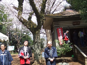 桜の木が立つ境内のような場所で、黒い羽織に白い花柄と赤い裏地が特徴的な半纏を着た数人の男性と、丸い屋根を持つ木造の古びたお堂を捉えた集合写真