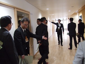 ビジネススーツを着た複数の男性が、薄い木目調の壁と床の明るい廊下で、一人の男性が前方へ腕を伸ばして説明をしながら歩いている様子の写真