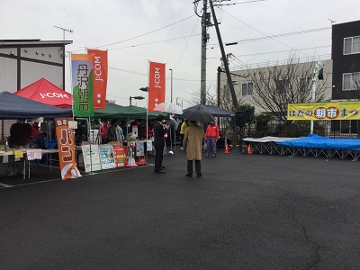 黒いアスファルトの広場に旗が立ち、テントや舞台が設置され、雨の降る曇り空の下で数人が集まっているはだの朝市まつりの様子の写真