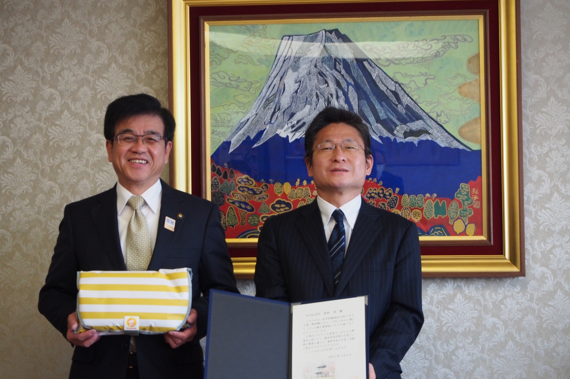 富士山の絵画が飾られた部屋で、市長は黄色と白の包み、男性は書類を持って共に笑顔で並んでいる写真