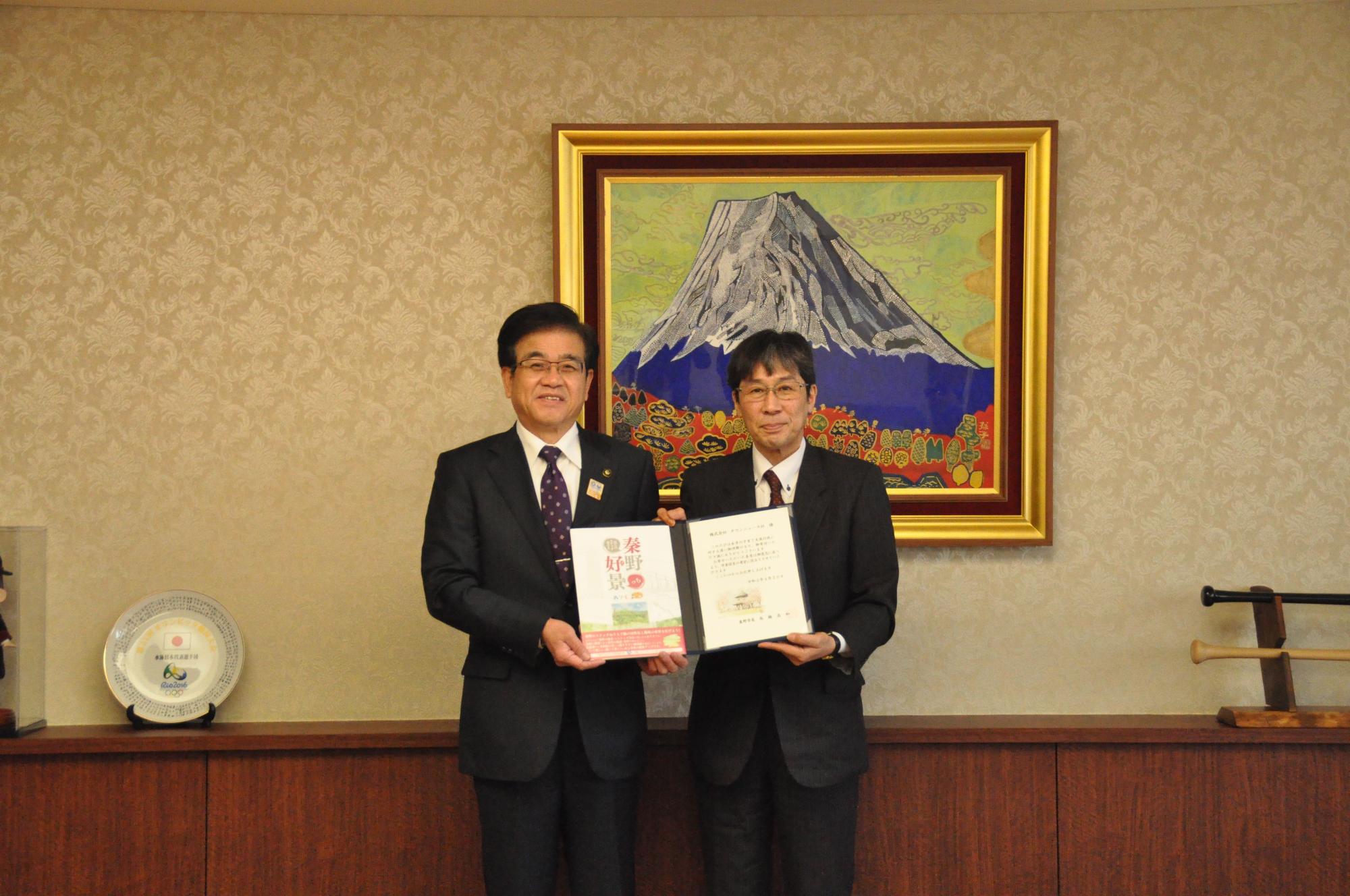 富士山の絵画が飾られた部屋で、市長と男性が笑顔で資料を持って並んでいる写真