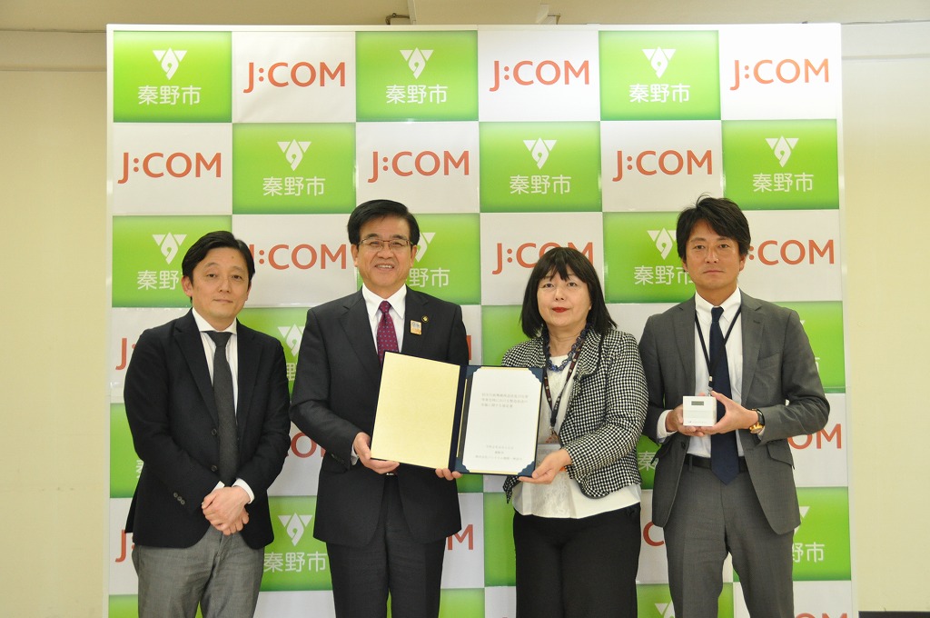 秦野市のロゴとJ:COMのロゴが並んだ背景ボードの前で、市長が女性と一緒に協定書を持ち、その両脇の男性の一人は白い箱を持って記念撮影をしている写真
