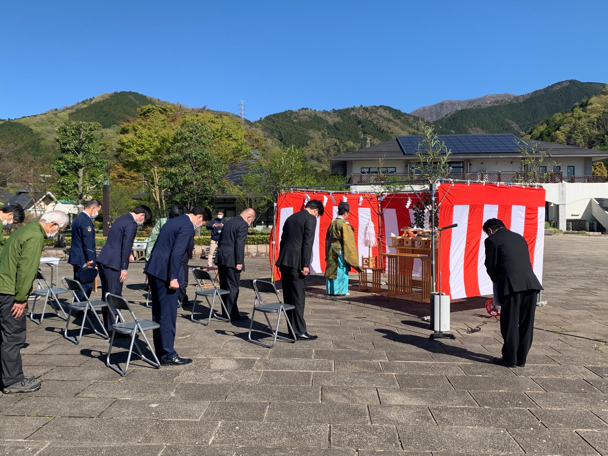 快晴の青空の下、スーツ姿の複数の参列者たちが、赤と白の紅白幕で設営された祭壇に向かって深く一礼をしている、地鎮祭の様子を写した写真
