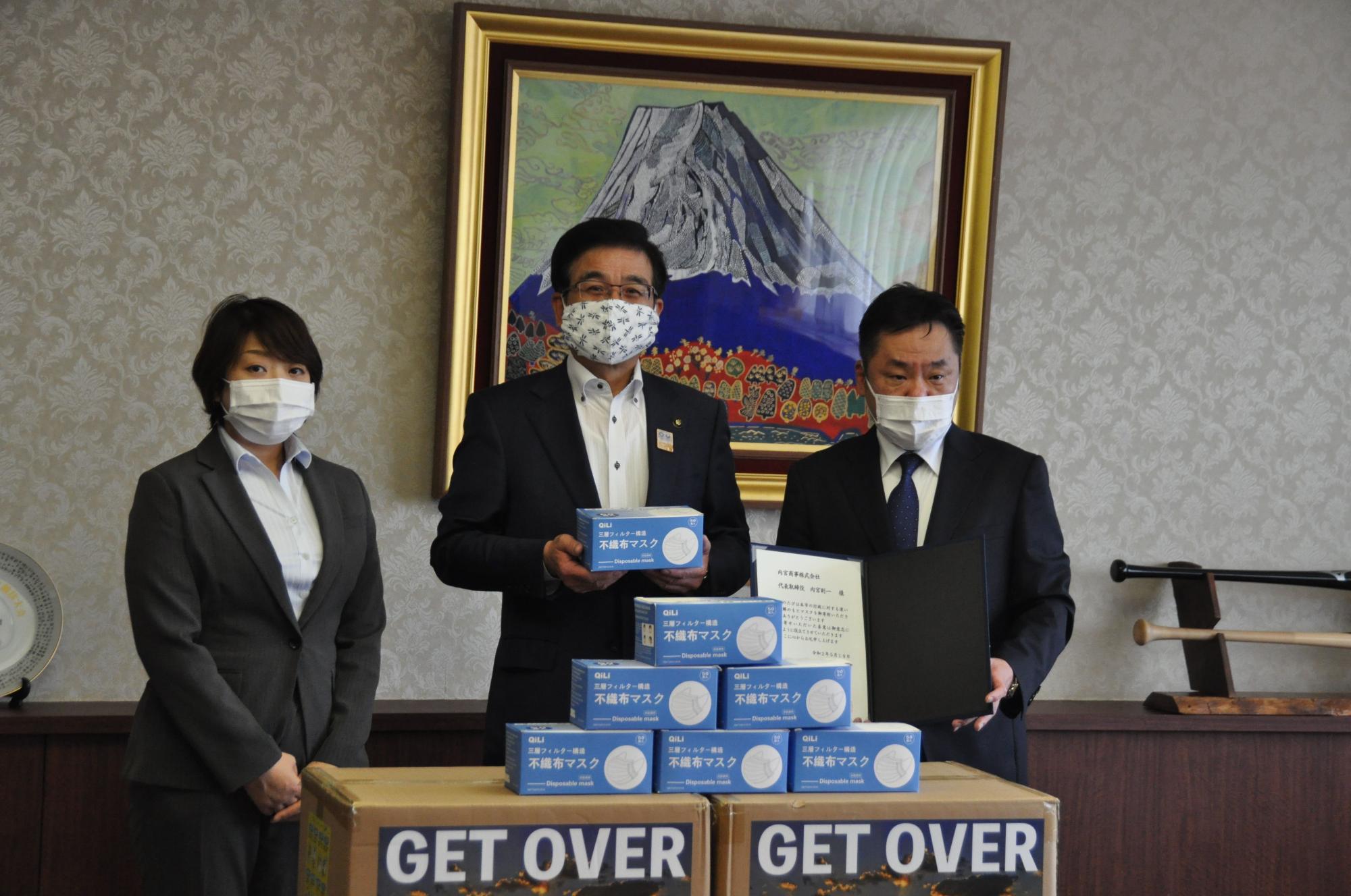 富士山の絵画が飾られた部屋で、マスクを着用した市長が青いマスクの箱、両脇の男女2人の男性が感謝状を持ち、段ボールを前にして立っている写真