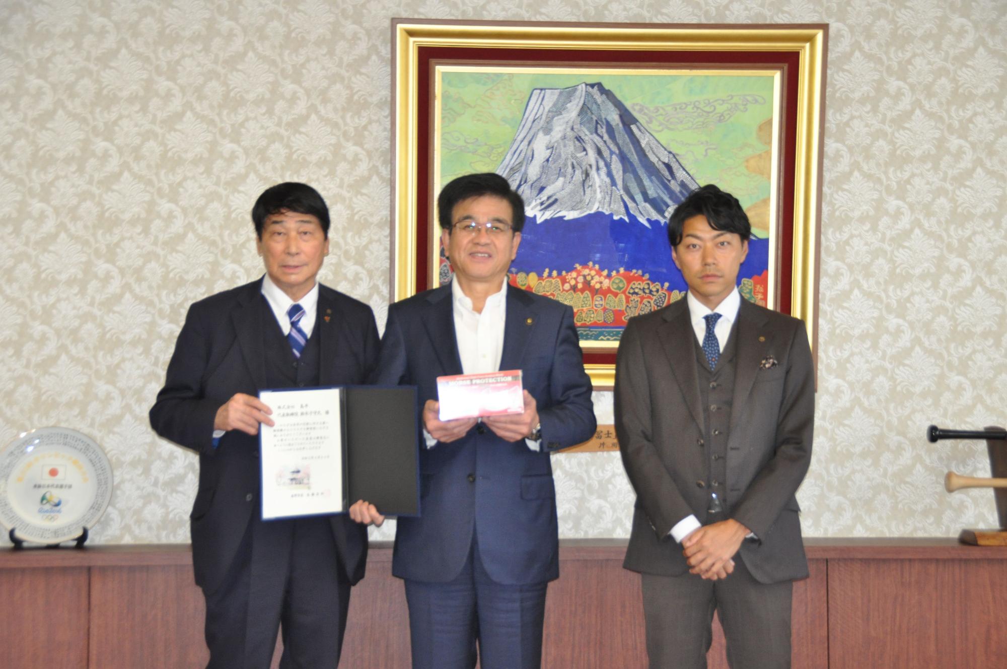 富士山の絵画が飾られた部屋で、3人のスーツ姿の真ん中の市長は白い箱のようなものを、左の男性は書類を持って並んでいる写真