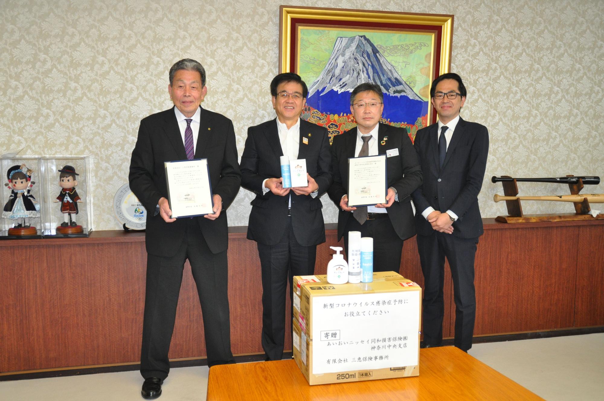 富士山の絵画が飾られた部屋で、スーツ姿の市長と3人の男性が、除菌スプレーなどの入った寄贈品の箱と、感謝状を持って並んでいる写真