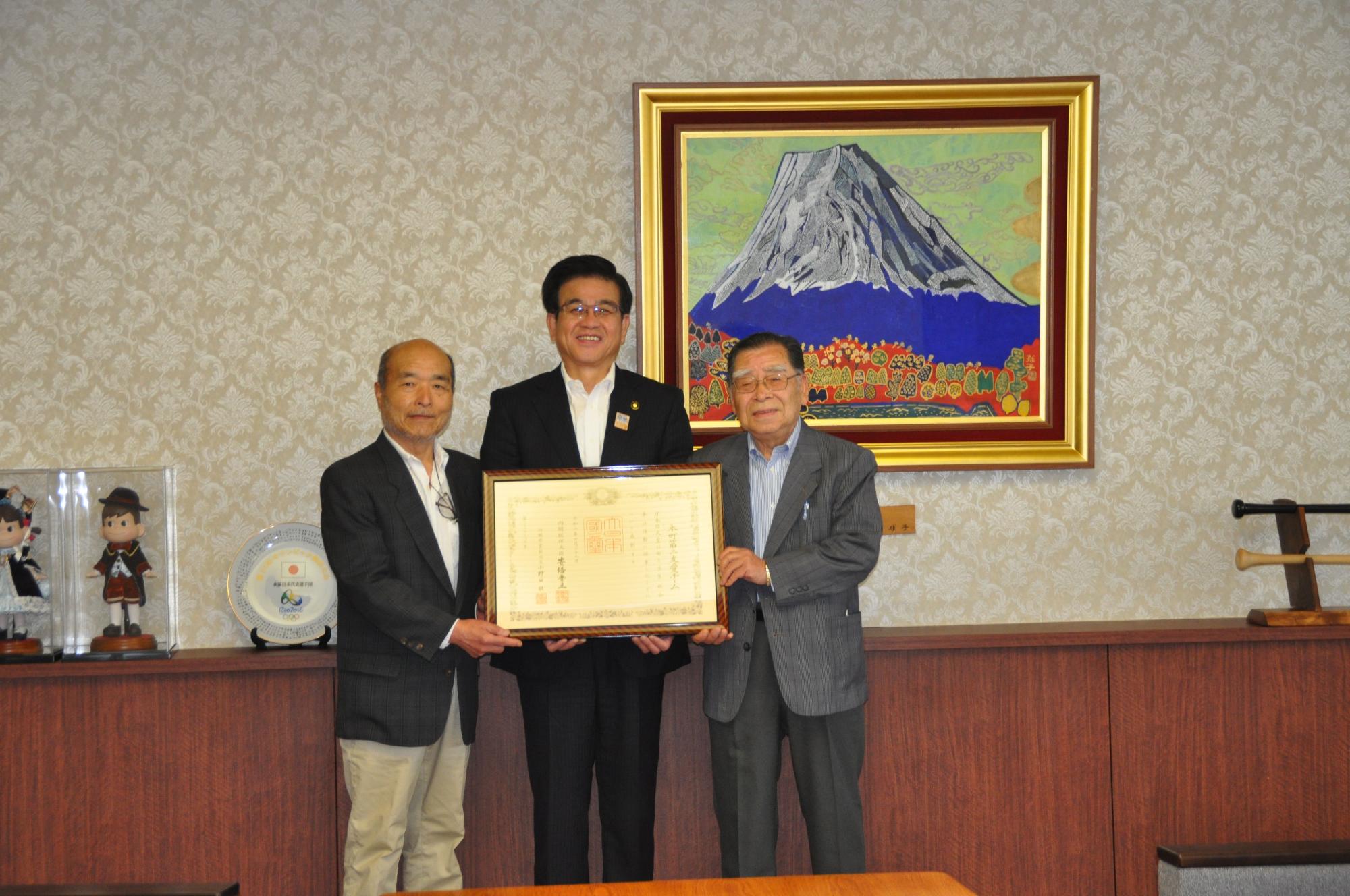 富士山の絵画が飾られた部屋で、市長の両脇に男性が立ち、3人で額縁に入った賞状を持って、記念撮影をしている写真