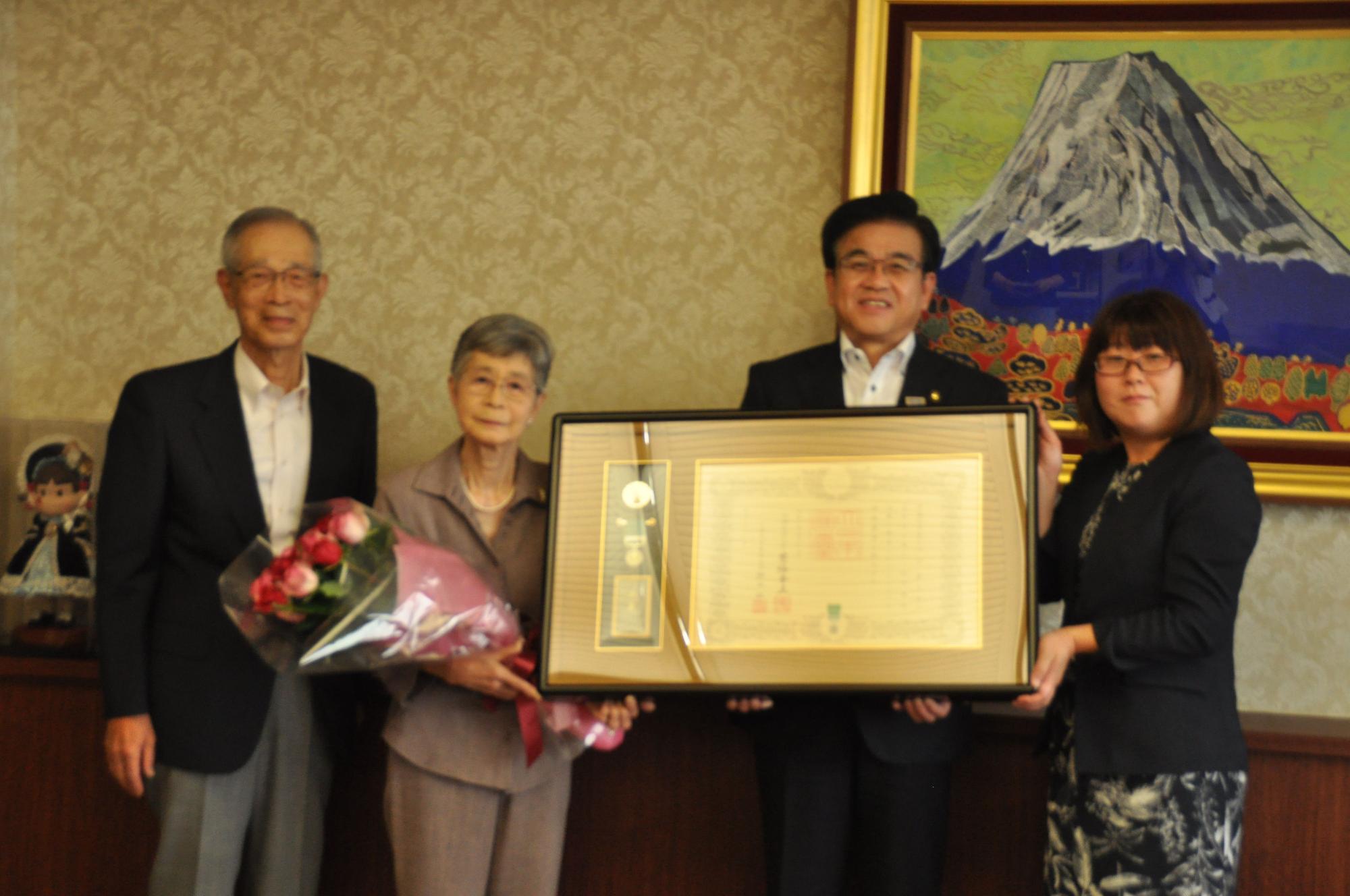 富士山の絵画が飾られた部屋で、年配の男性、花束を持った山田喜久子さん、大きな額縁に入った賞状を市長と女性で持ち、計4人が並んで写っている記念写真