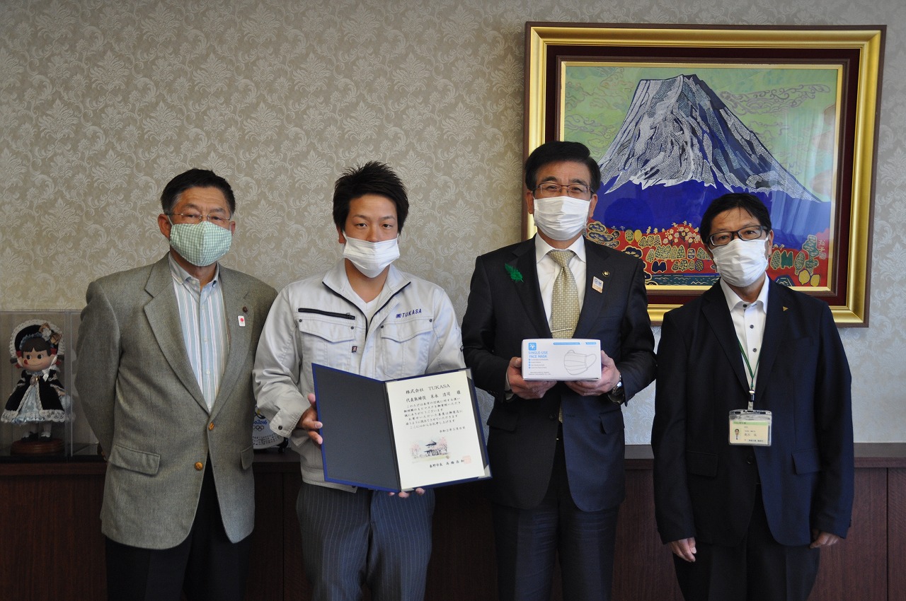 富士山の絵画を背景に、マスクを着用した市長と3人の男性が、感謝状とマスクの箱を持って並んでいる寄贈式典の写真