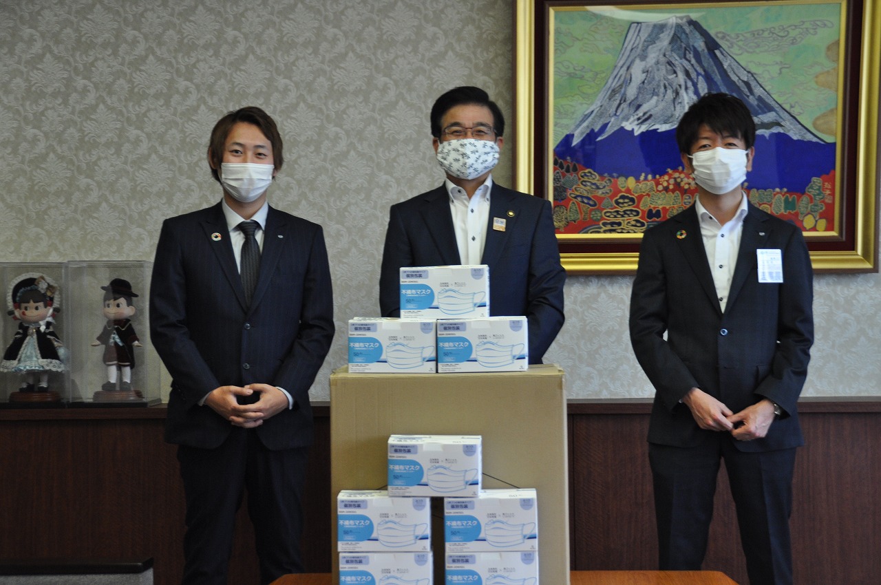 富士山の絵画を背景に、マスクを着用し、スーツ姿の市長と2人の男性が、寄贈されたマスクの段ボール箱の前に並んで写っている写真