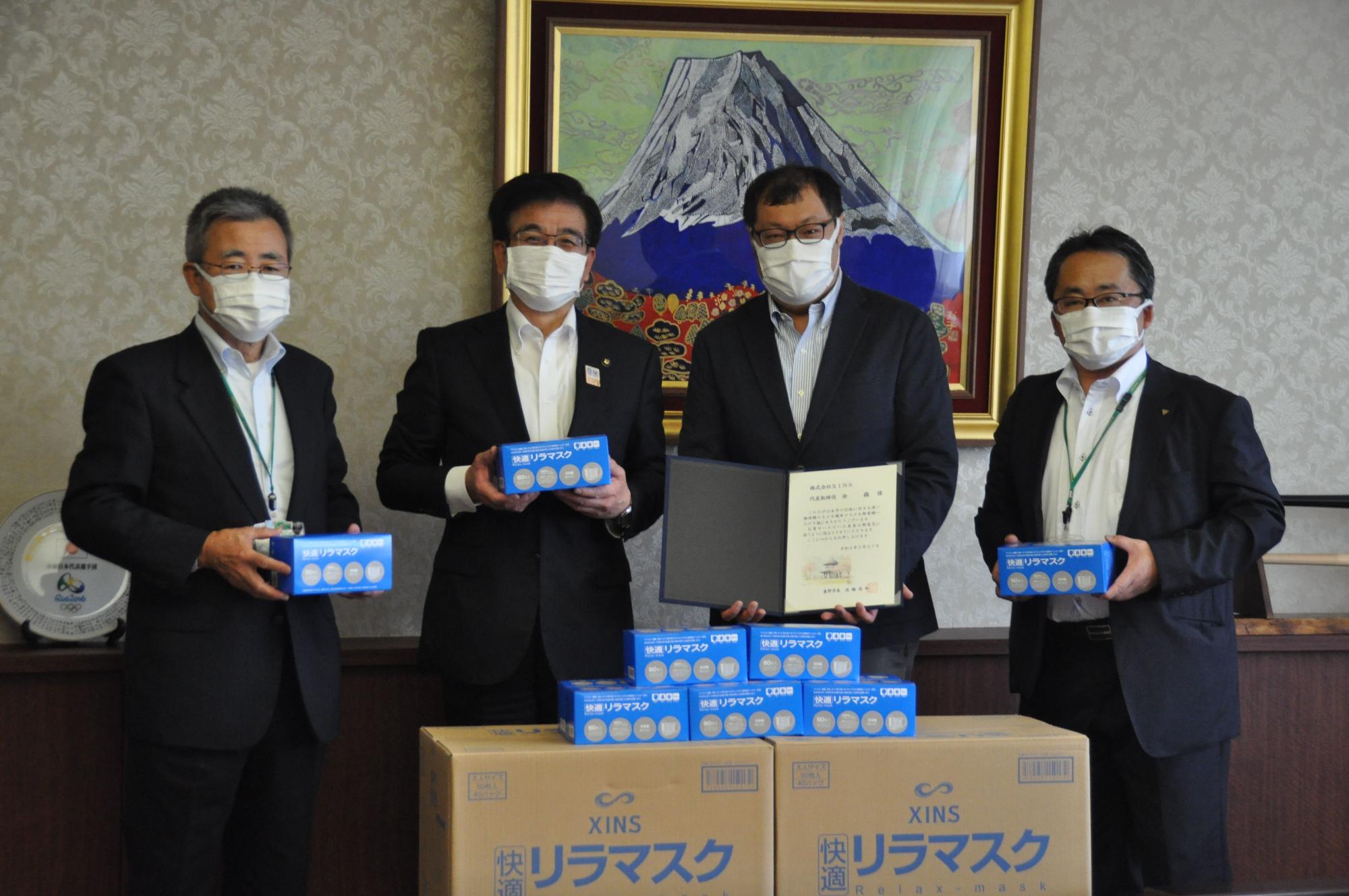富士山の絵画が飾られた部屋で、複数のマスクの箱と、協定書のような書類を囲んで、スーツ姿の市長と3人の男性がマスクを着用して立っている写真