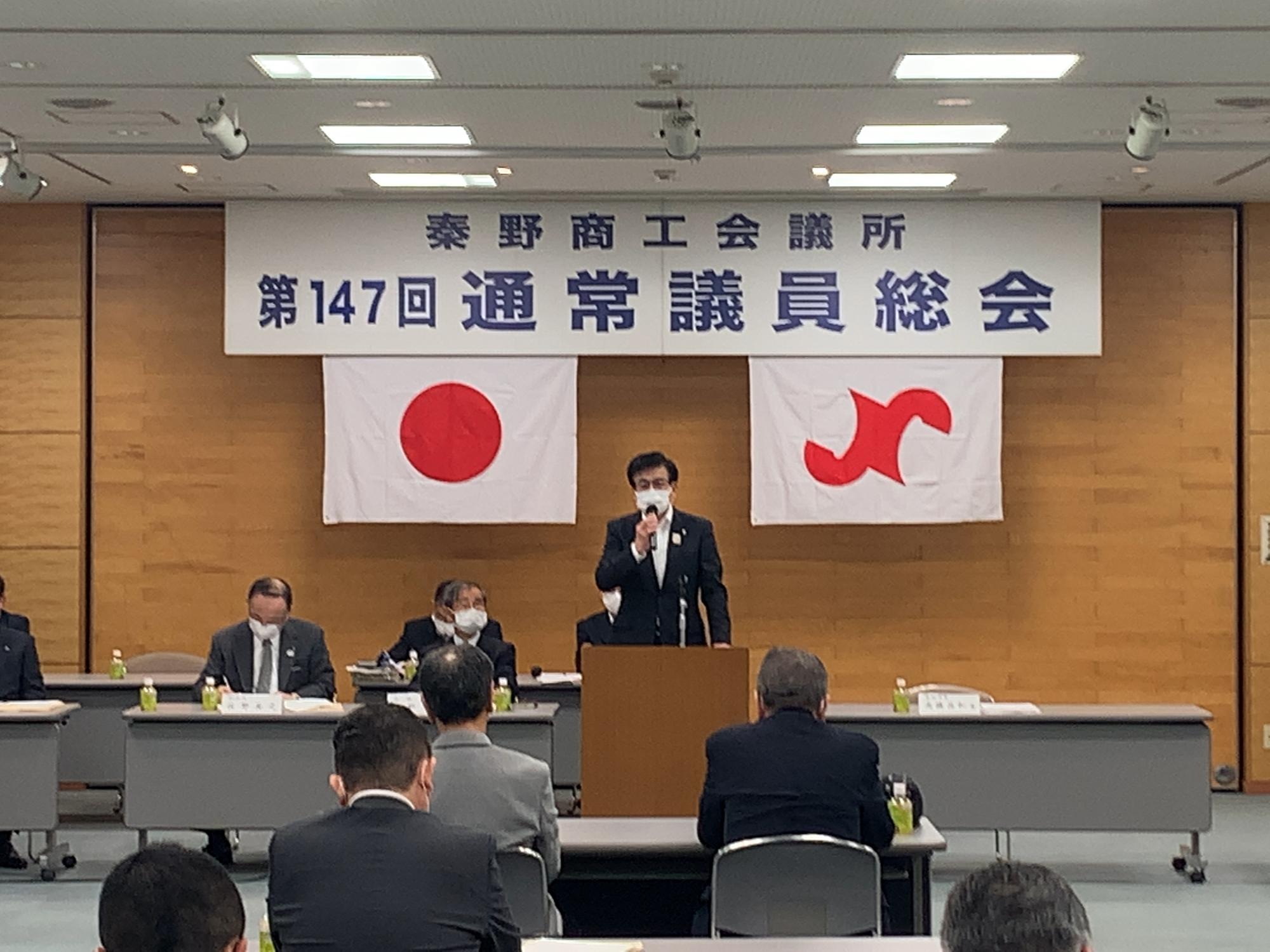 「秦野商工会議所 第147回通常議員総会」の横断幕が掲げられた会場で、マイクを持ったスーツ姿の市長が、多くの議員を前に挨拶をしている写真