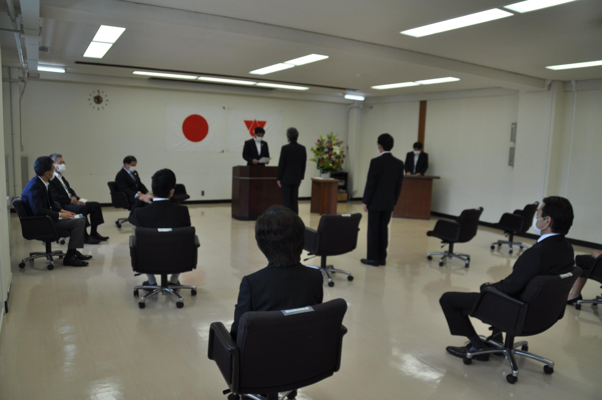 日本国旗と市旗が掲げられた部屋で、マスクを着用した複数の参加者が間隔を空けて着席し、壇上の人物が挨拶をしている写真