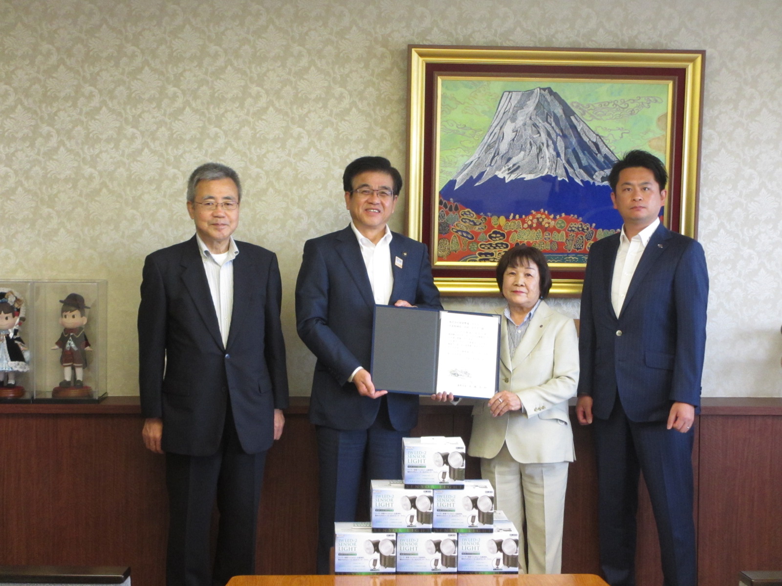 富士山の絵画が飾られた室内で、スーツ姿の男性3人と女性1人が並んでおり、中央の市長と女性が感謝状を持ち、手前には記念品のセンサーライトの箱が置かれている写真