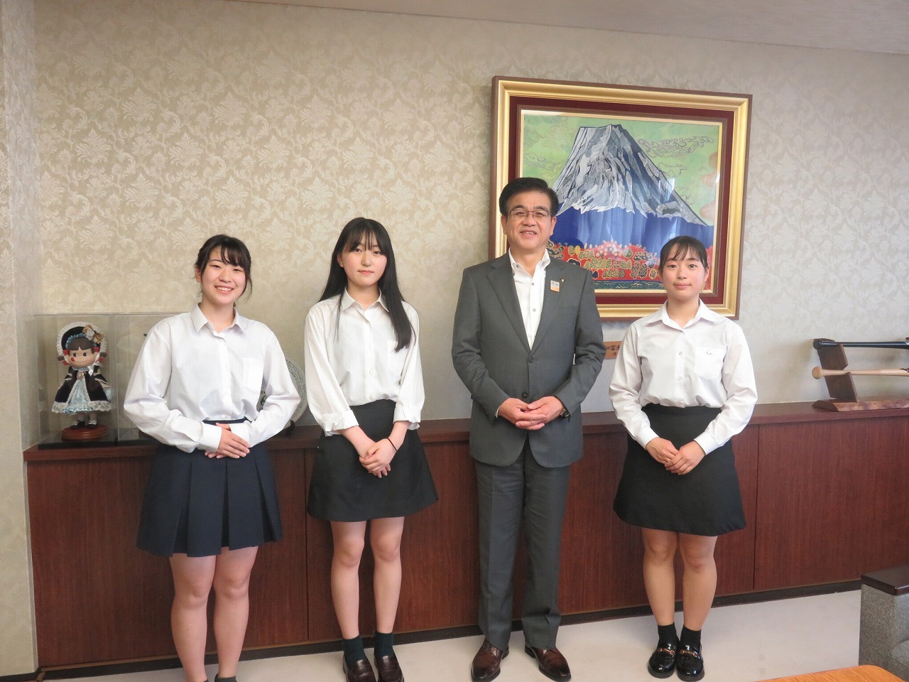 スーツ姿の市長を囲んで制服姿の女子学生3人が並んでおり、全員が両手を前でくんでカメラに微笑んでいる集合写真