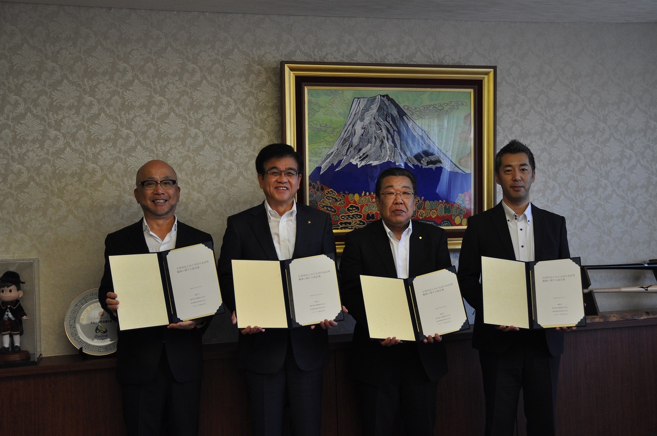 背景に富士山の絵画が飾られている室内で、市長と3人のスーツ姿の男性が、それぞれ協定書を両手で持ってカメラに向かって笑顔で並んでいる写真