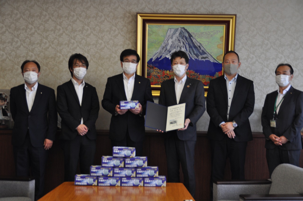 富士山の絵画が飾られた室内で、マスクを着用したスーツ姿の6人の男性の中で、市長がマスクの箱、隣の男性が感謝状を持ち、手前には積み上げられたマスクの箱が並んでいる写真