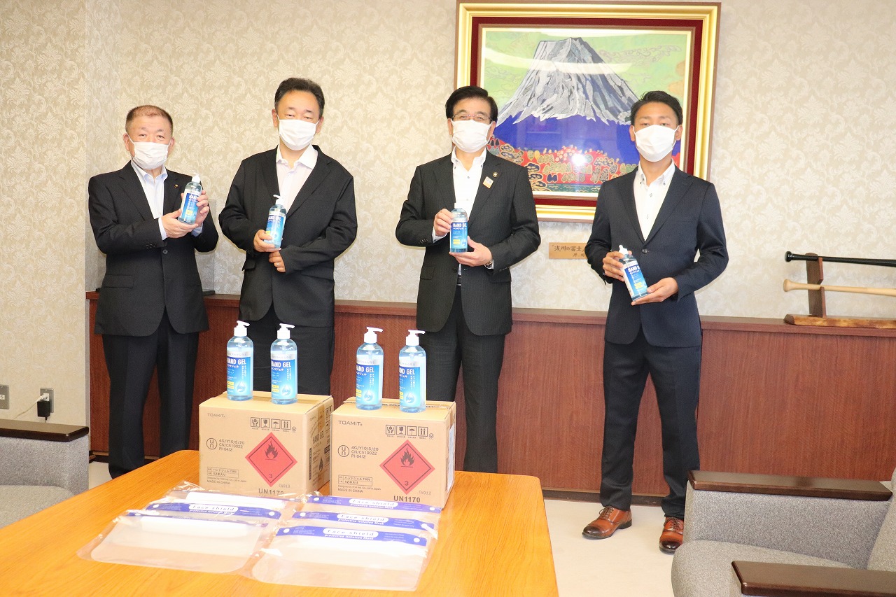 富士山の絵画が飾られた室内で、マスクをつけた市長と3人の男性がアルコール消毒液を手に持ち、手前には段ボール箱やフェイスシールドなどの寄贈品が並んでいる写真