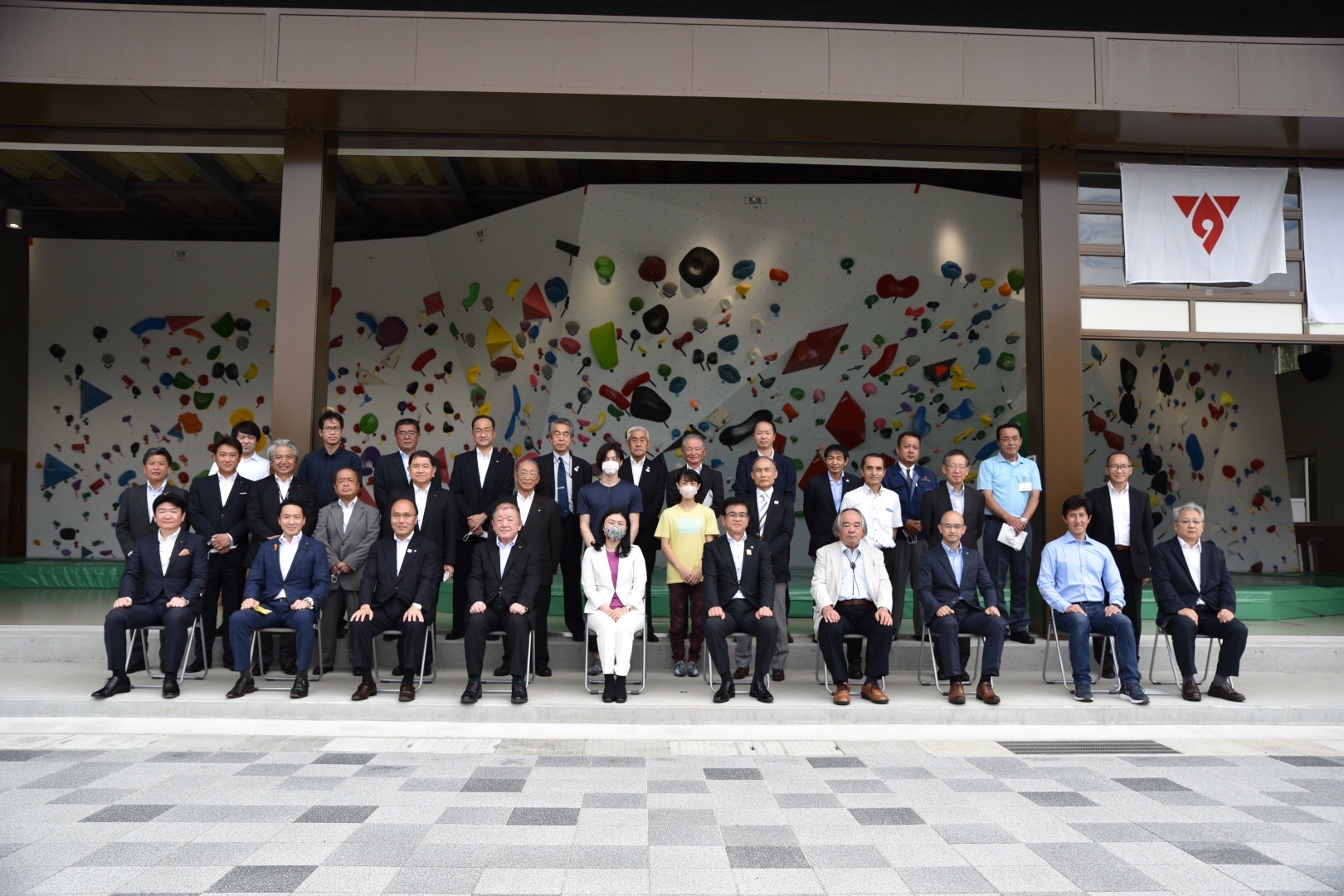 色とりどりのホールドが設置されたボルダリングウォールを背景に、多くの人がスーツ姿で並んでおり、前列の10名は椅子に座り、他の方々は2列に並んでいる写真