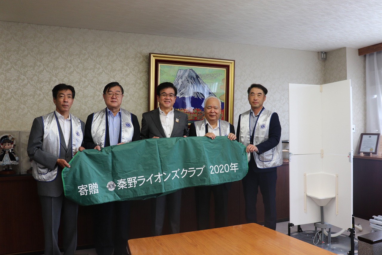 富士山の絵画が飾られた室内で、市長と4人の男性が緑色の大きな布を持ち、組み立て式の簡易トイレを挟んで並んでいる写真