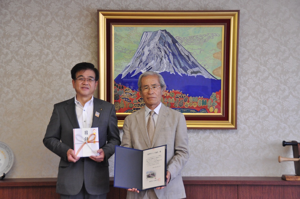 富士山の絵画が飾られている室内で、左側のスーツ姿の市長が目録を持ち、右側の男性が感謝状を持っている並んでいる写真