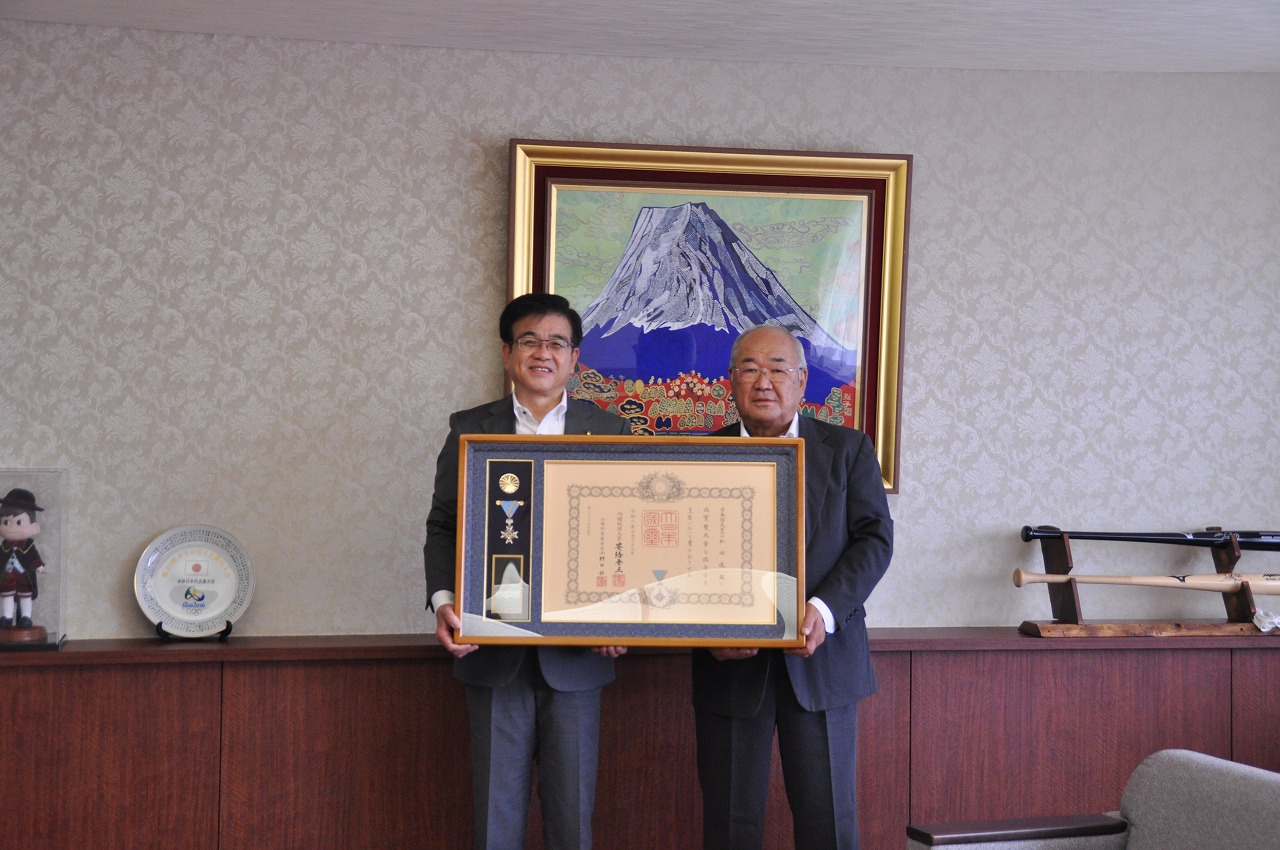 富士山の絵画が飾られた室内で、スーツ姿の市長と男性が勲章と賞状の入った額縁を両手で持ち、笑顔で並んでいる写真