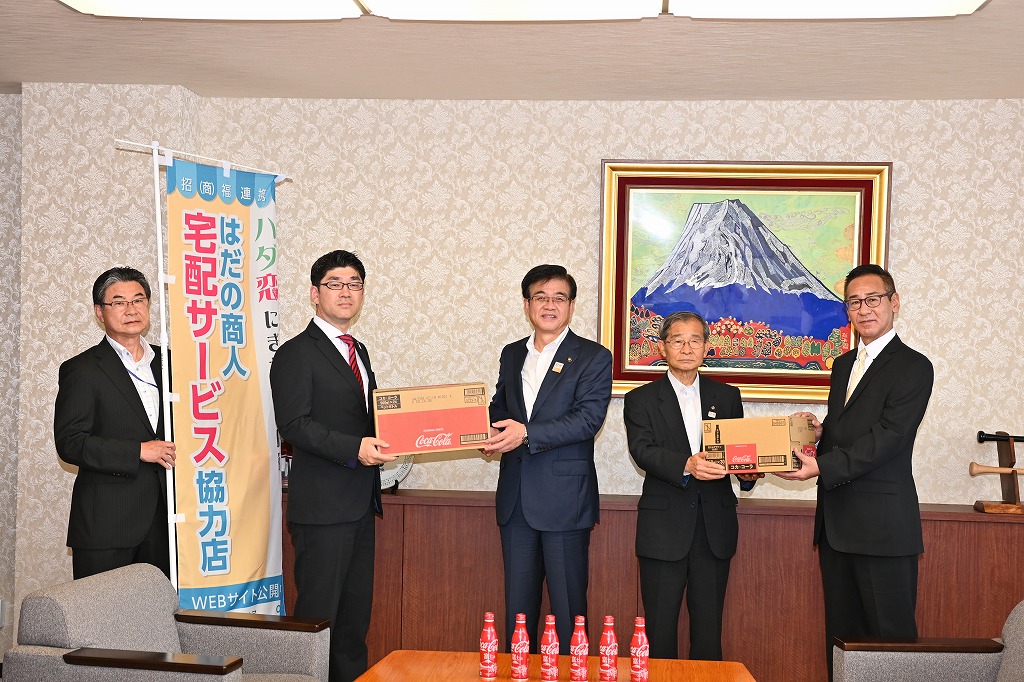 富士山の絵画が飾られた部屋で、スーツ姿の市長と4人の男性が、飲み物の箱や幟を持って並んでいる、贈呈式の写真