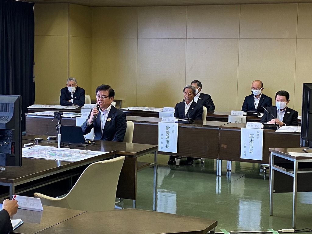 マスクを着用したスーツ姿の男性たちが、他の市長などの役職名が書かれた名札を立てて、会議室で長机を囲み、秦野市長がマイクで発言している写真