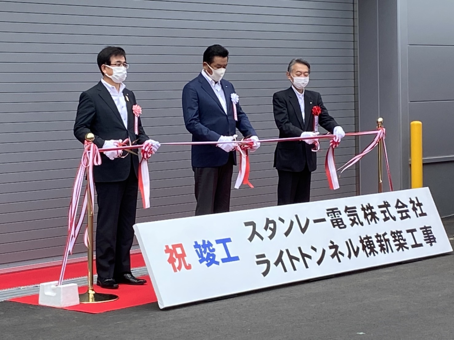 「祝 竣工 スタンレー電気株式会社 ライトトンネル棟新築工事」と書かれた看板が手前に置かれた屋外で、スーツ姿の市長と他二人の男性が、紅白のテープにハサミを入れている様子を写した写真