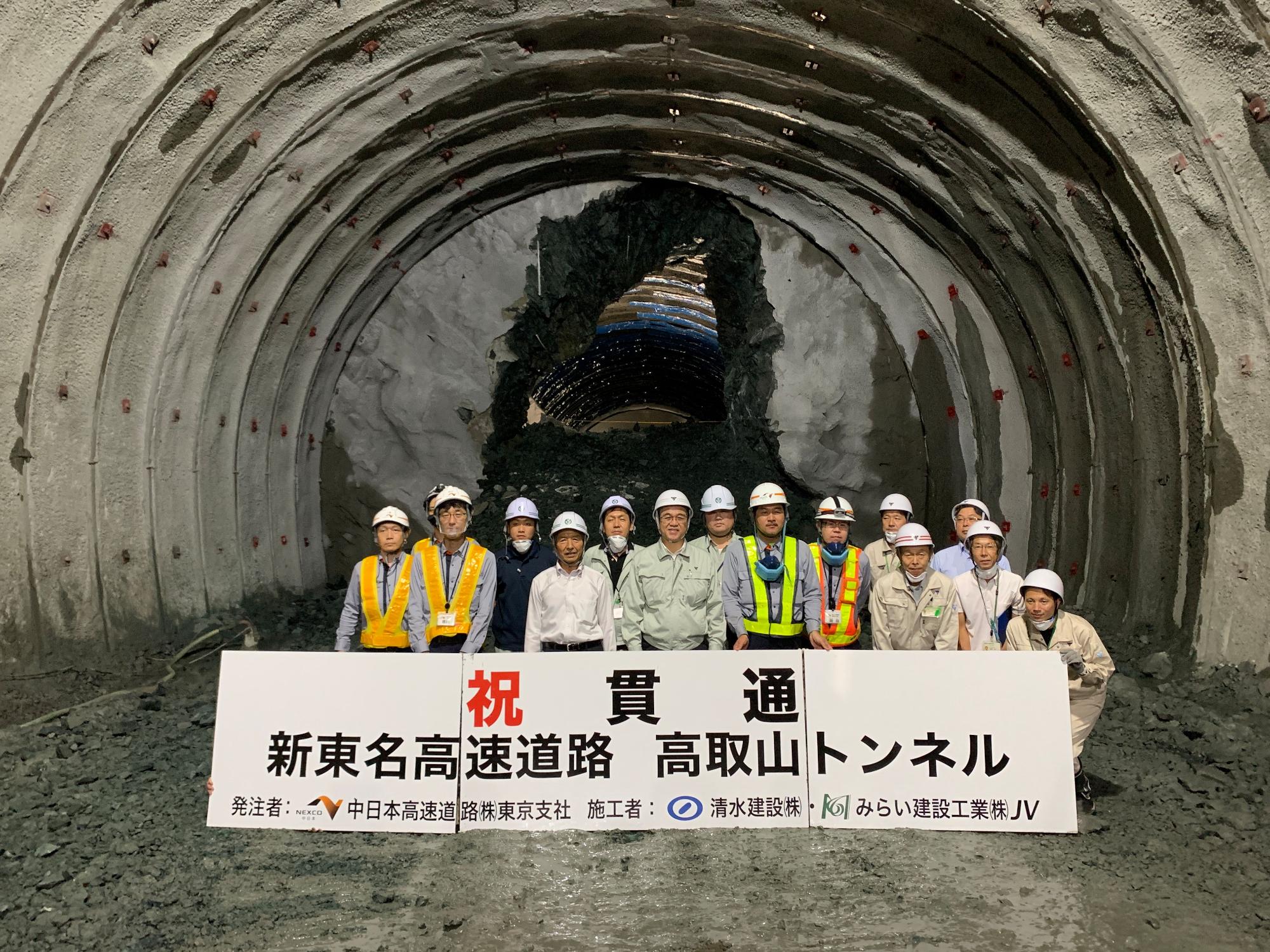 完成前のトンネル内部で、ヘルメットを被った複数の建設作業員が、「祝 貫通 新東名高速道路 高取山トンネル」と書かれた横断幕を持って写っている記念写真