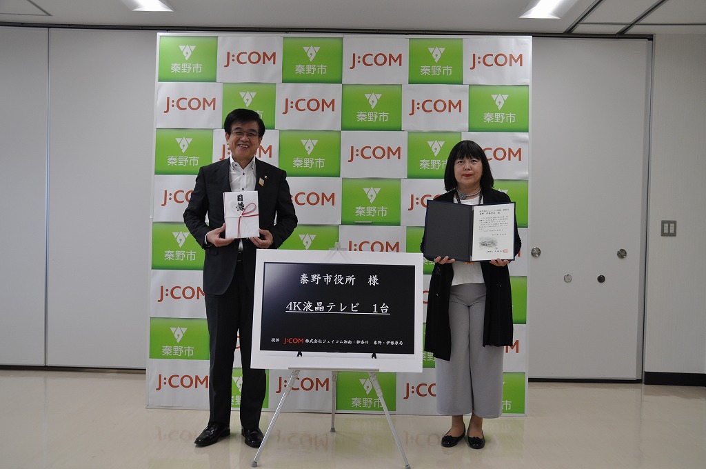 秦野市とJ:COMのロゴがデザインされた背景ボードの前で、市長は目録のようなもの、女性は感謝状のような書類を持ち、中央には「4K液晶テレビ 1台」と書かれたボードが置かれている写真
