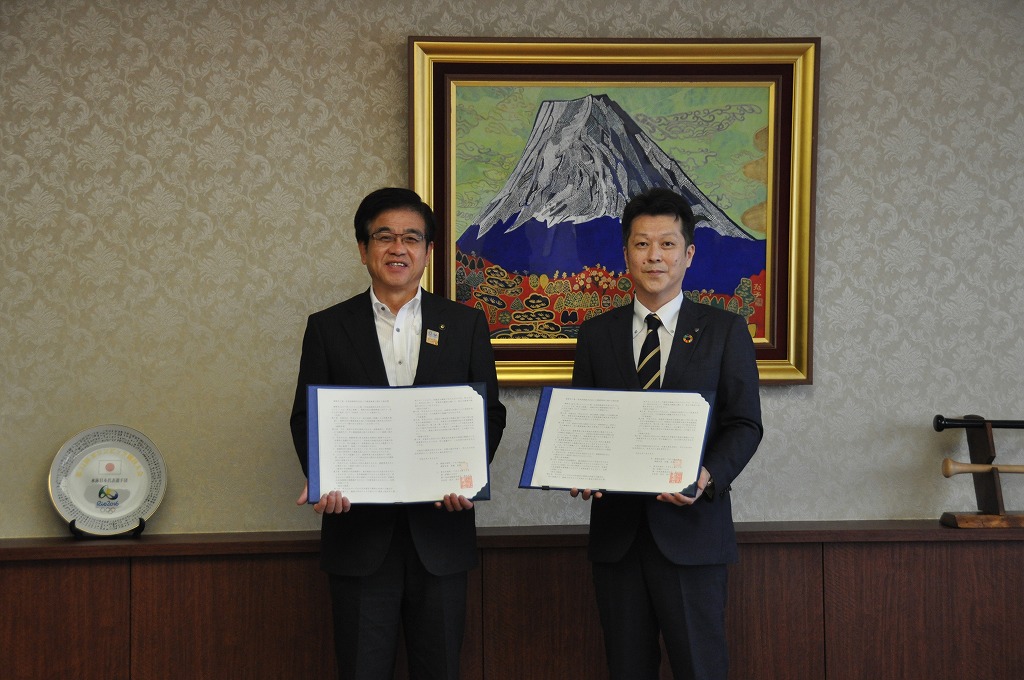 富士山の絵画が飾られた部屋で、スーツ姿の市長と男性が、それぞれ協定書を見開いて書類を持って立っている写真
