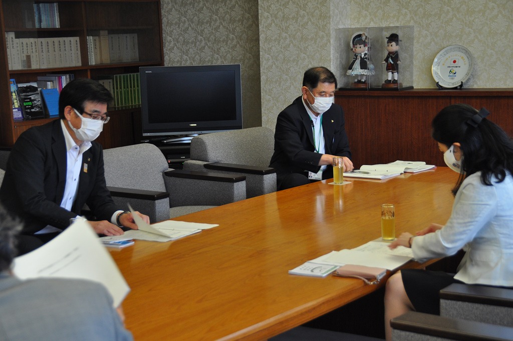 会議室のような部屋で、マスクを着用した市長や女性、他の複数の人が、大きな木製のテーブルを囲んで書類を見ながら会議をしている写真