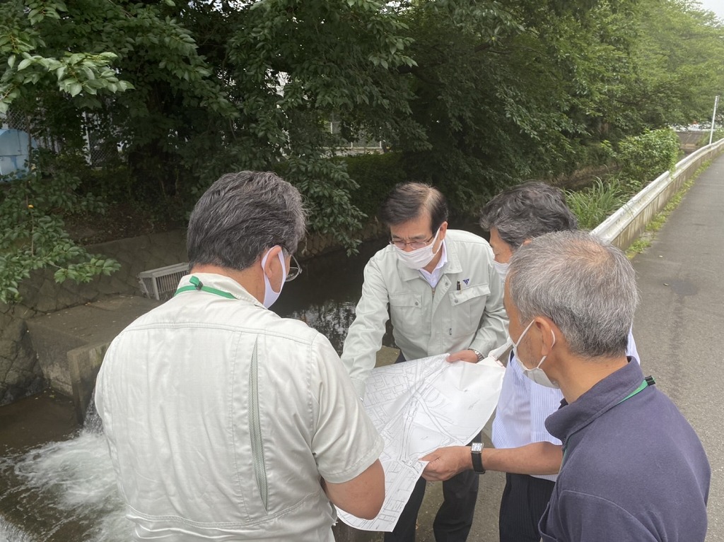 水が流れる水路の脇で、白いマスクを着用した市長と3人の男性が、広げた図面を見ながら話し合っている写真